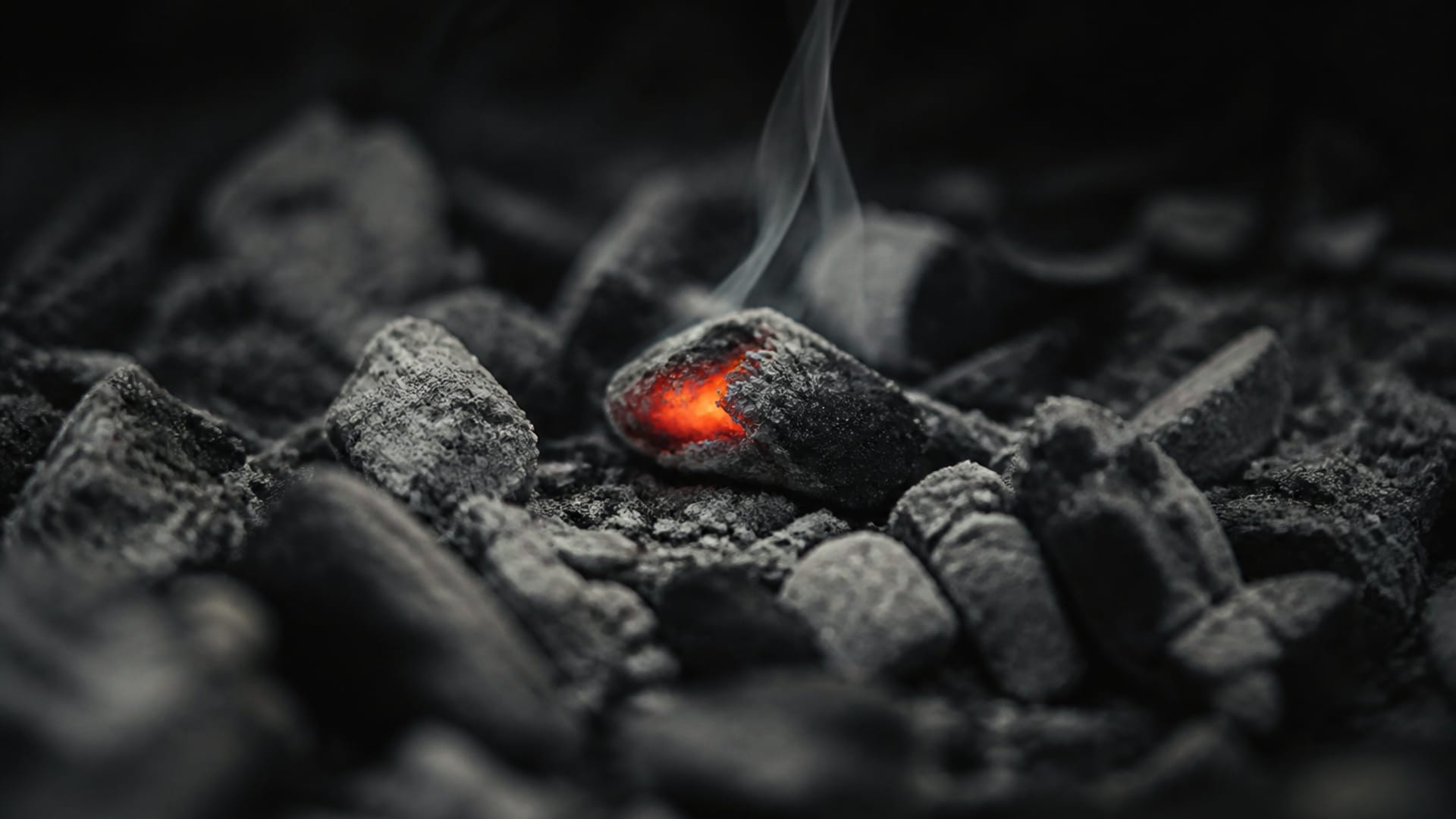 Uma única brasa viva acesa entre cinzas cinzentas de madeira, com um fio de fumaça subindo.