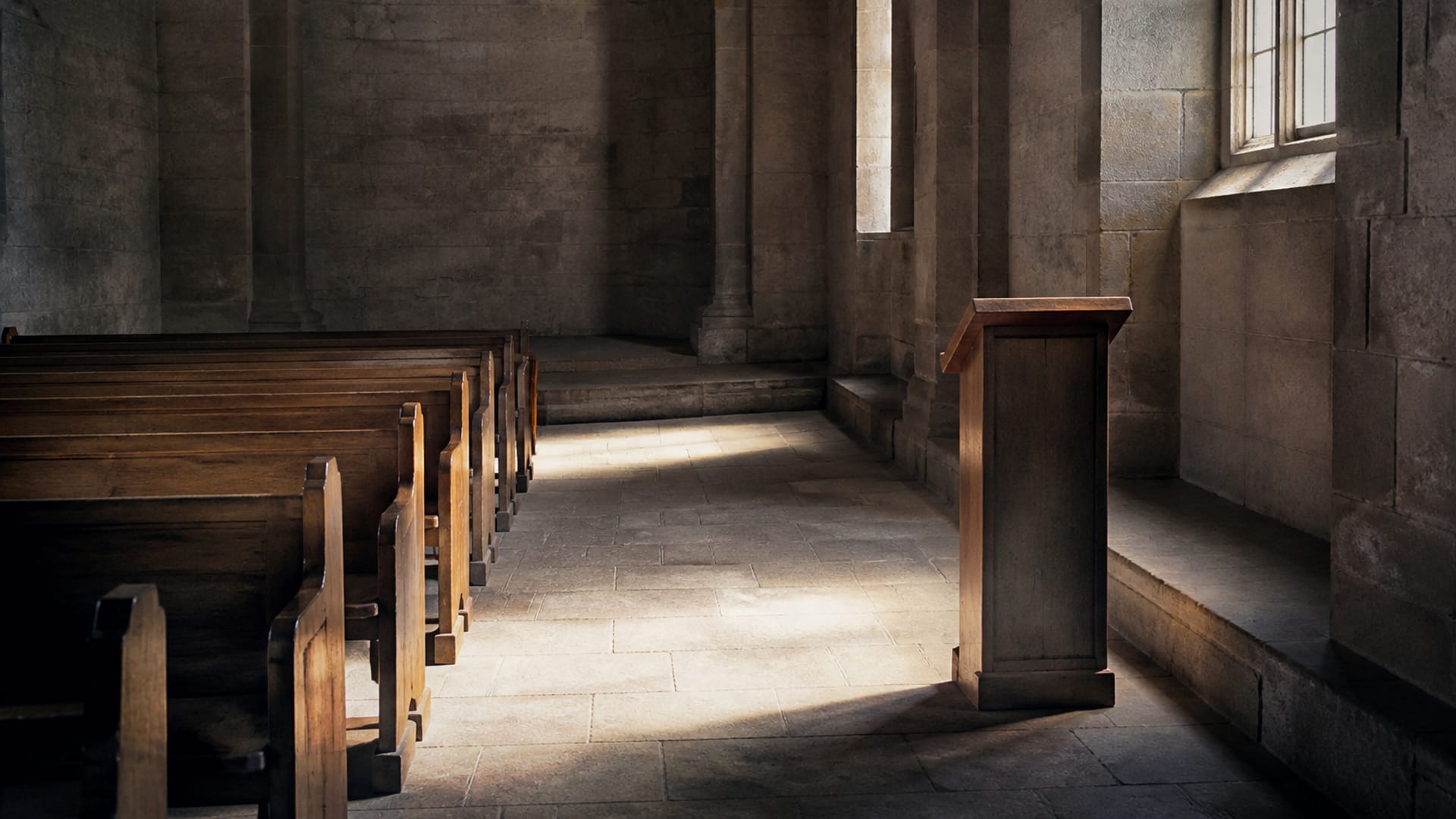 The inside of a plain stone church early in the morning, empty pews and a simple wooden lectern.