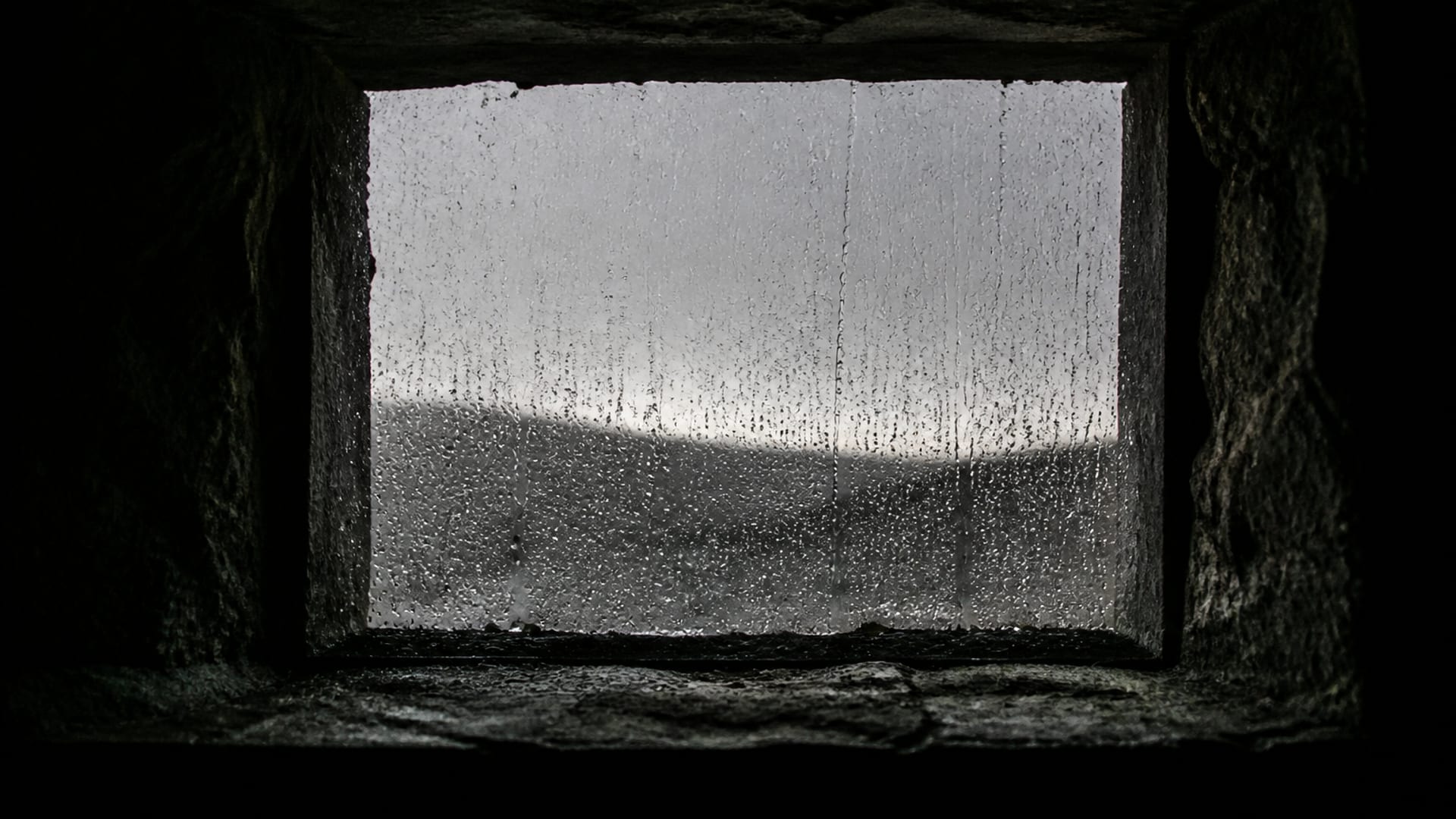Chuva escorrendo pelo lado de fora de uma pequena janela de moldura de pedra, vista de um cômodo escuro.