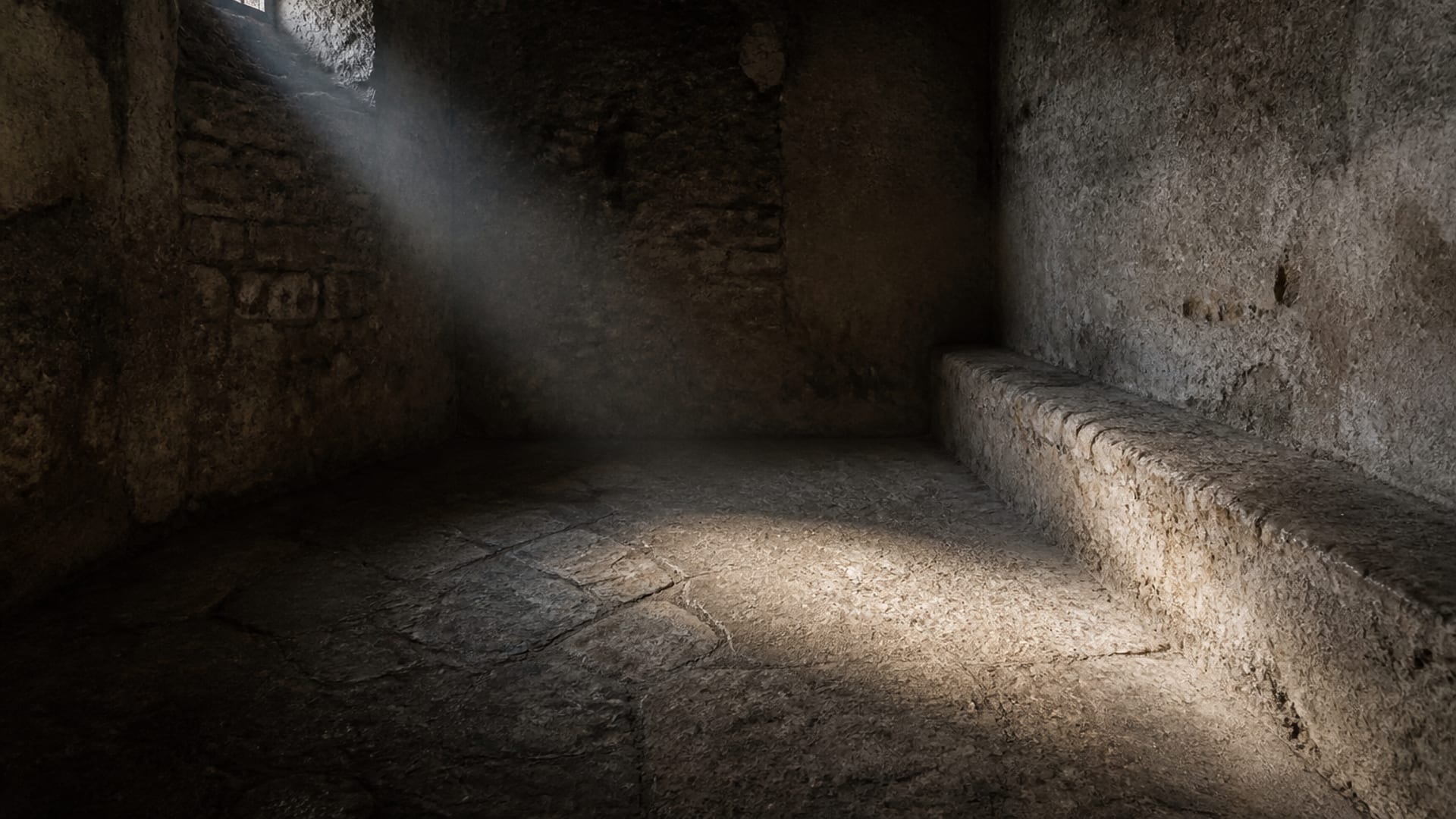 O interior de uma cela romana de pedra nua, com um único facho de luz vindo de uma abertura gradeada no alto e caindo sobre o chão gasto.