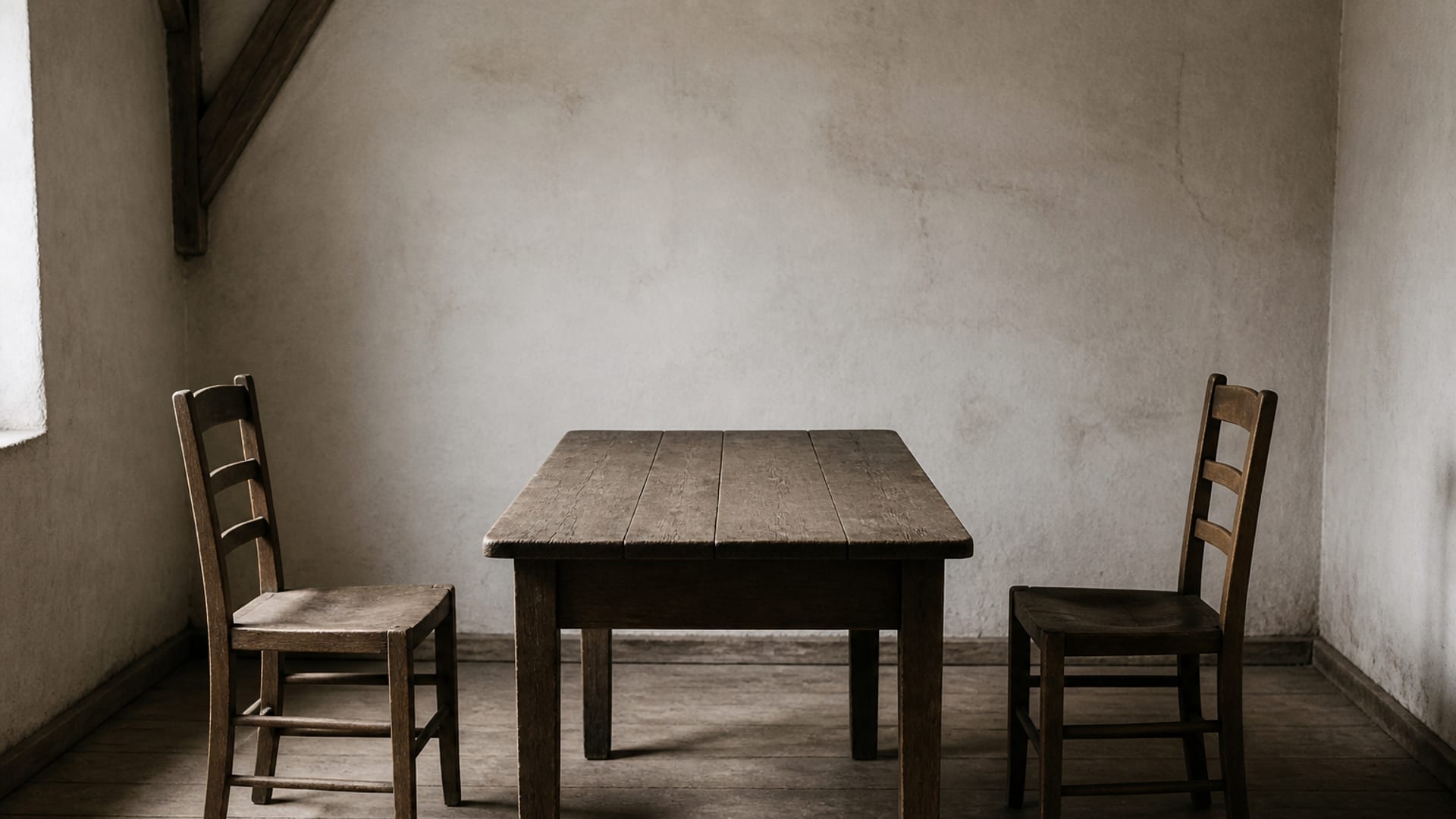 Two plain wooden chairs facing each other across a bare table in an empty whitewashed room.