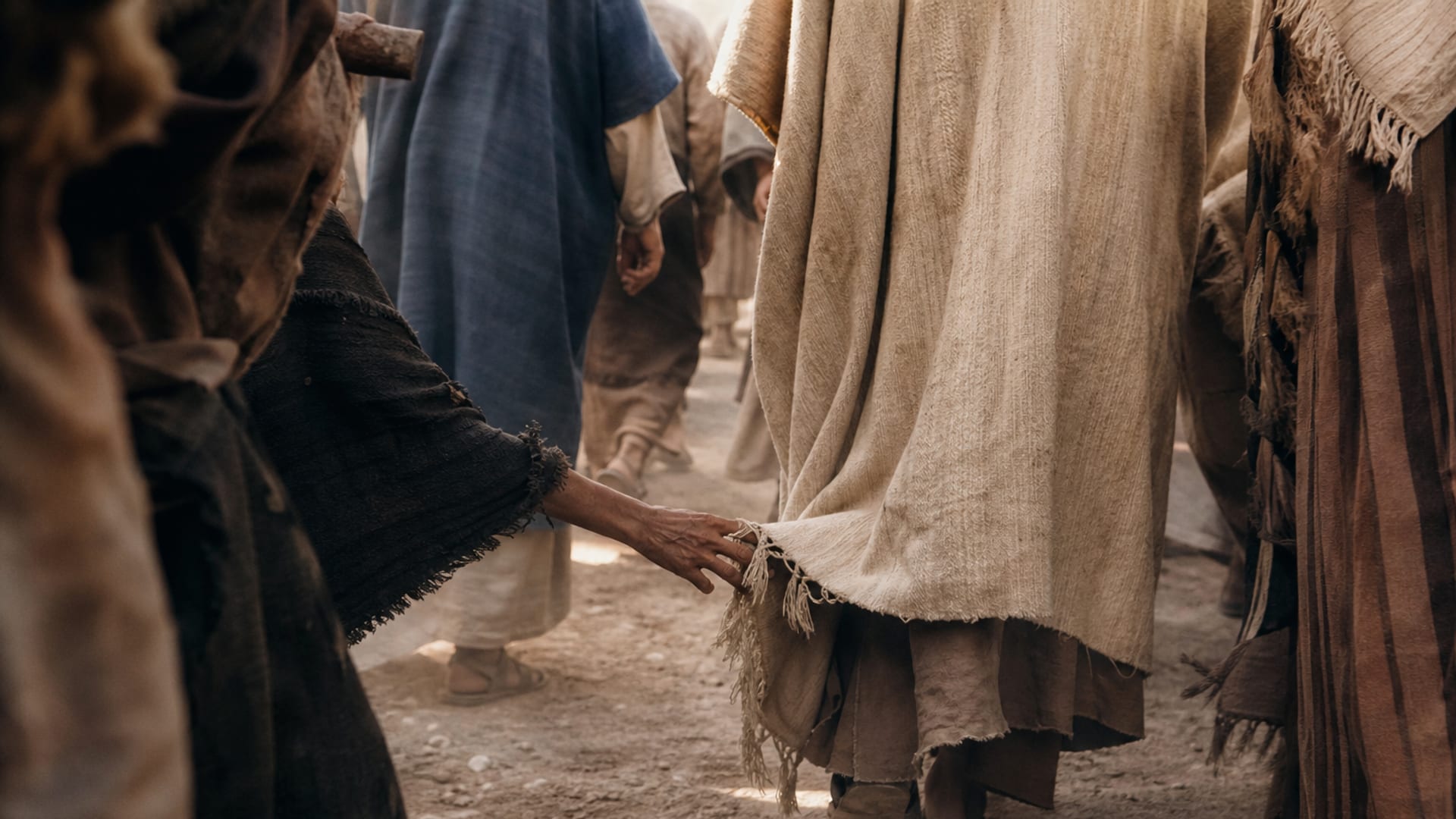 A close view in a crowded, dusty first-century lane: a woman's thin hand reaching between moving bodies toward the knotted fringe at the edge of a traveller's plain wool cloak.