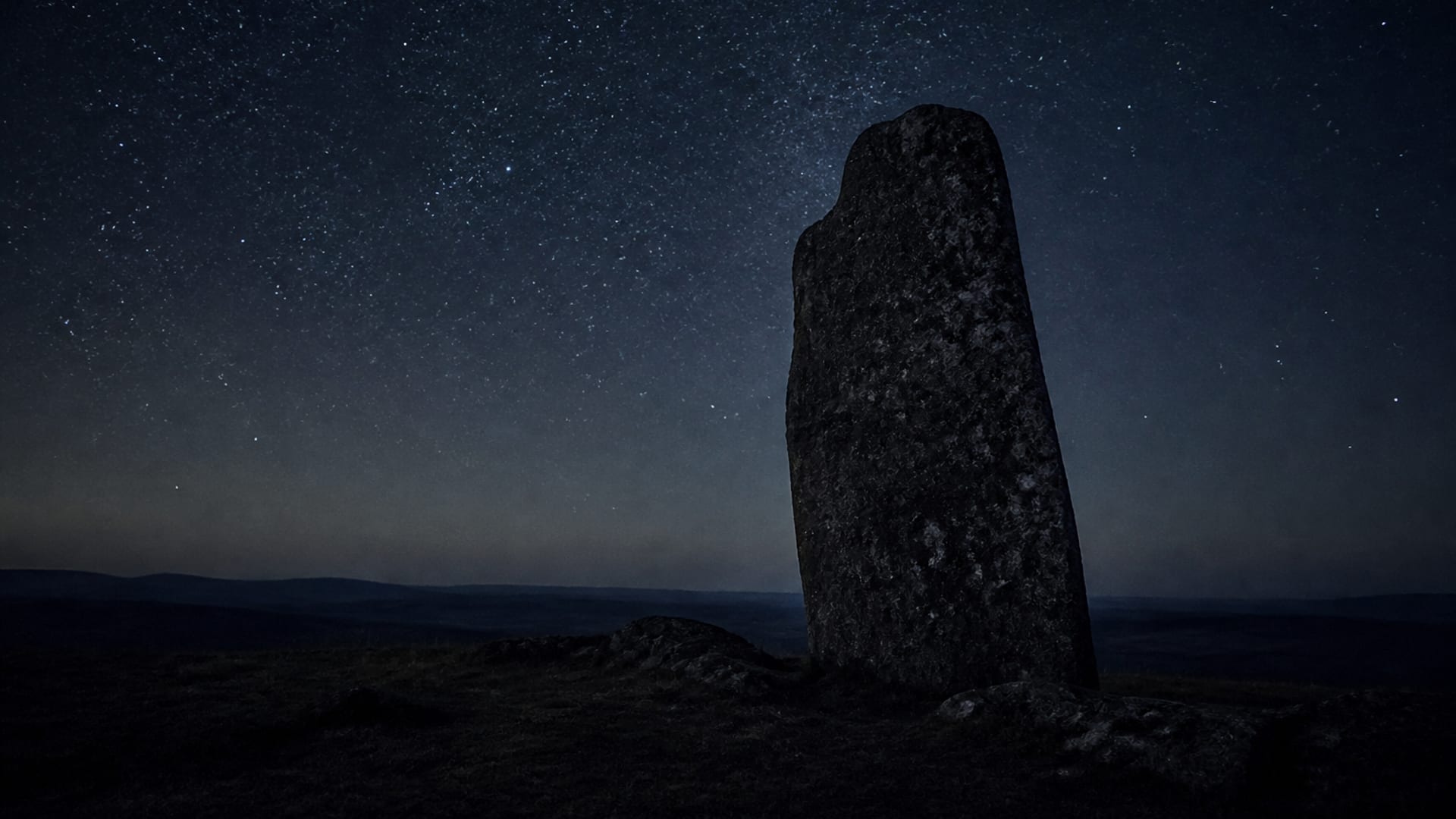 A single rough standing stone on bare hill country, silhouetted against a deep field of stars.