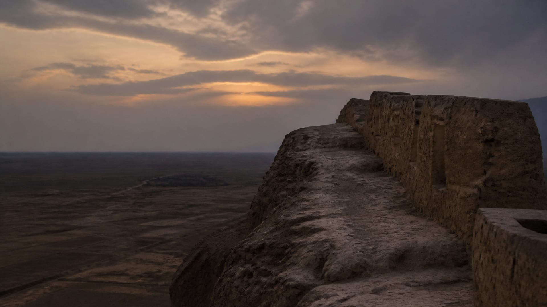 The top of an ancient mudbrick rampart at dusk, the parapet running away toward an empty plain.