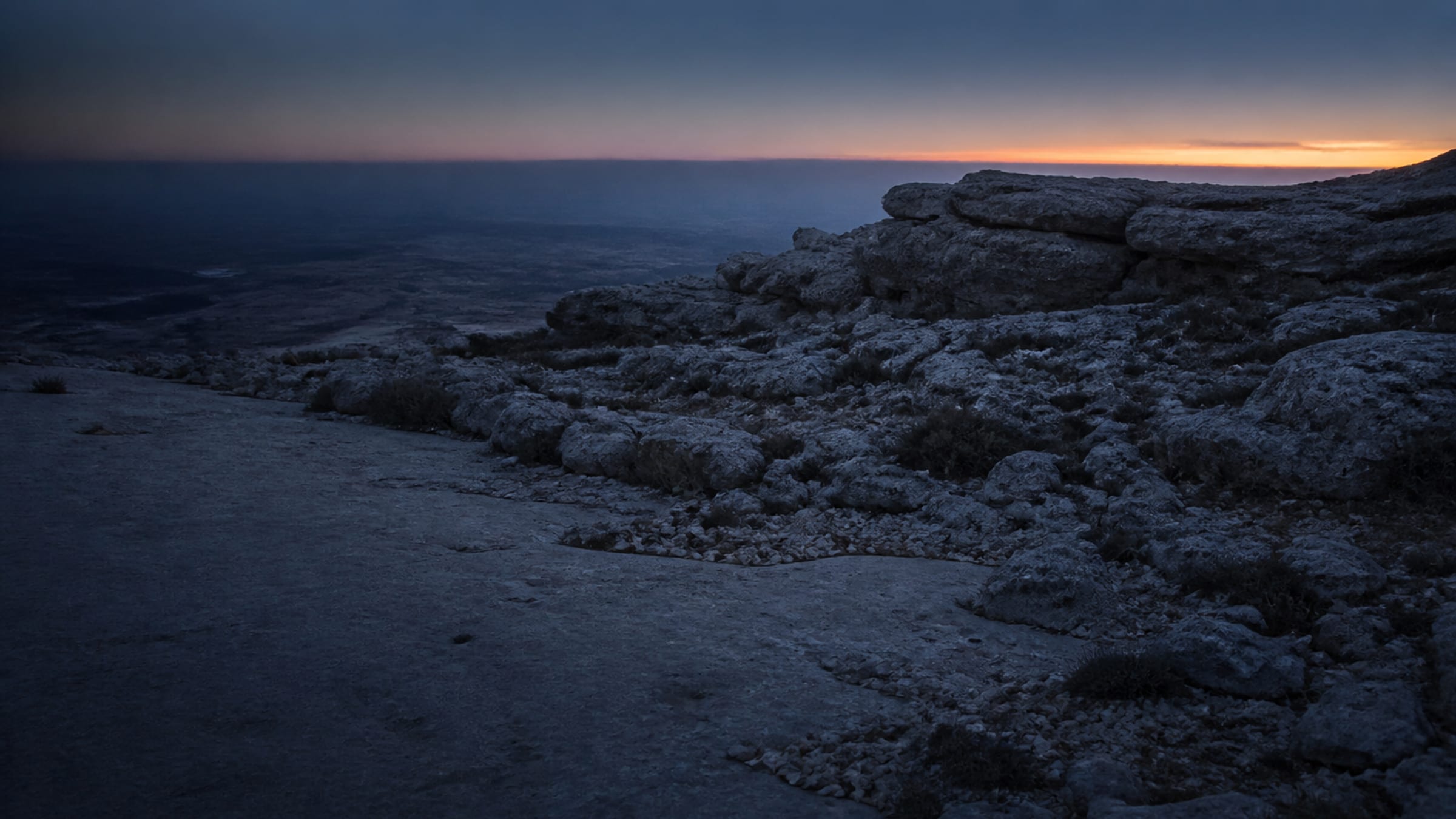 A rocky limestone hillside in the blue darkness before sunrise, with a thin band of warm light along the far horizon.