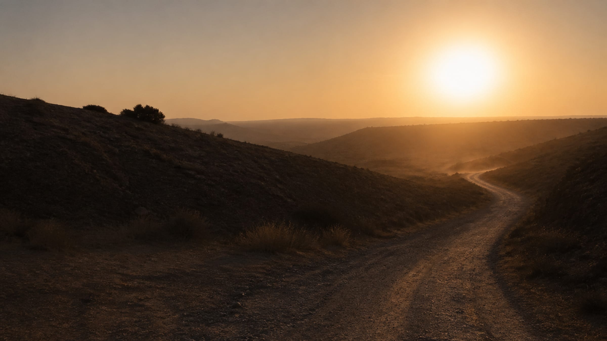 Uma estrada de terra fazendo curva entre colinas baixas ao entardecer, com poeira no ar e o sol perto do horizonte.