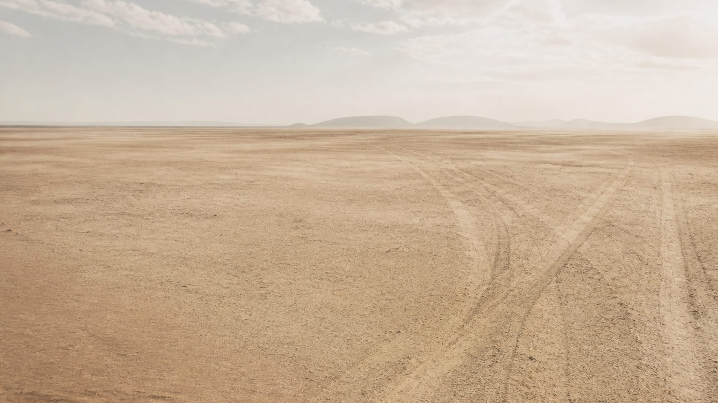 A wide desert plain in pale haze, with faint old tracks crossing the sand toward distant low hills.